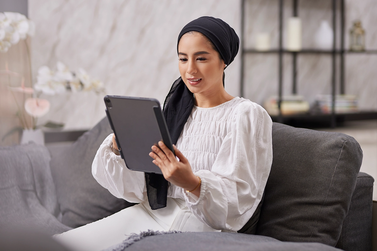 Woman in headscarf sitting on couch using tablet device in modern living room with natural lighting