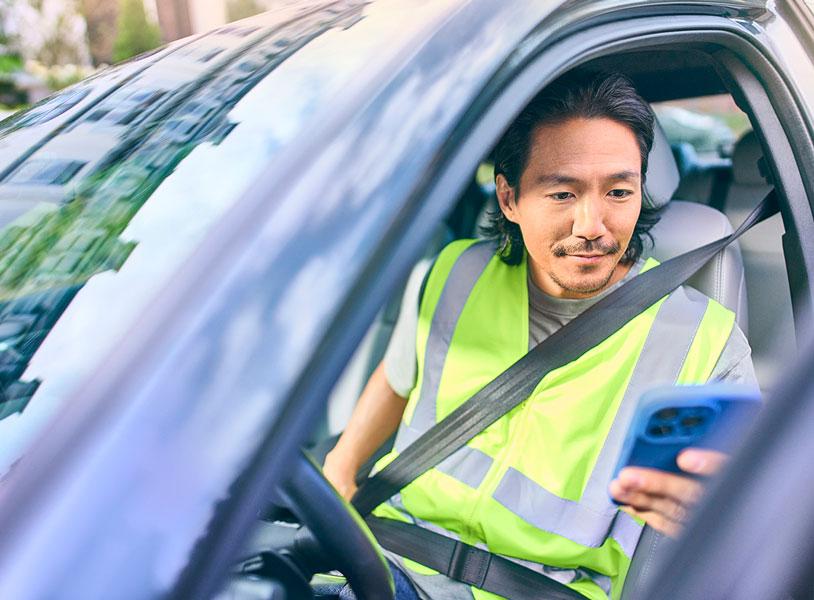 Delivery driver in safety vest seated in car holding smartphone while wearing seatbelt on residential street