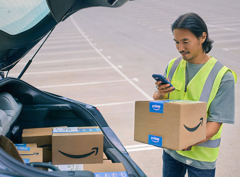  Delivery driver in safety vest holding Amazon Prime package while checking phone near open car trunk with more boxes
