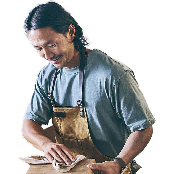 Man in blue t-shirt and tan apron smiling while wiping wooden cutting board with cloth with a yellow safety vest in the background