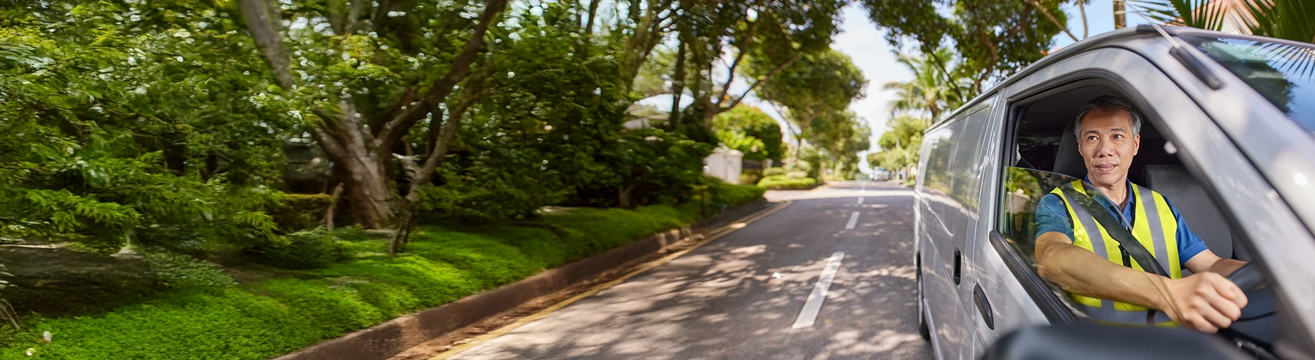 Delivery driver in high-visibility vest driving a van on tree-lined residential street