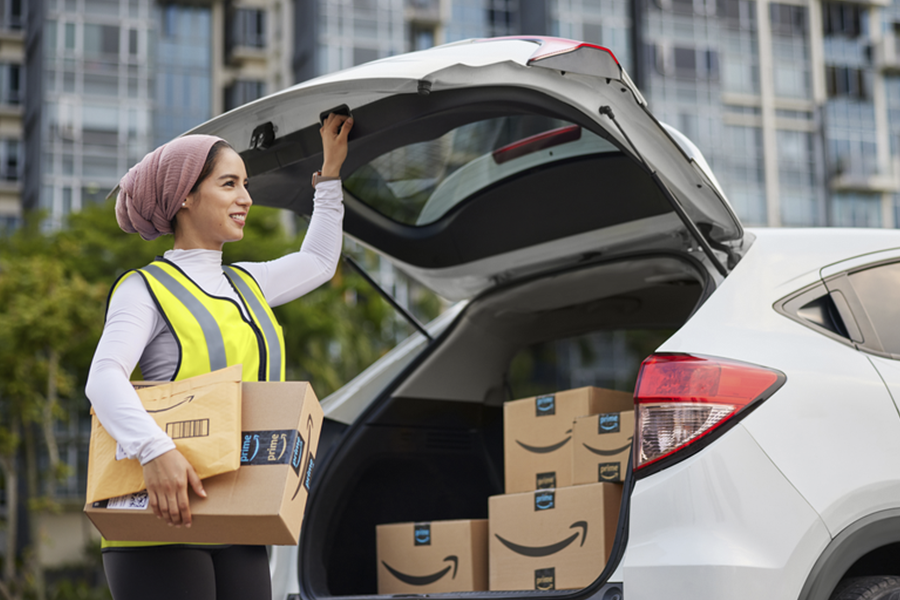 Woman in a yellow vest holding Amazon packages in one hand and closing the trunk of her car with the other.