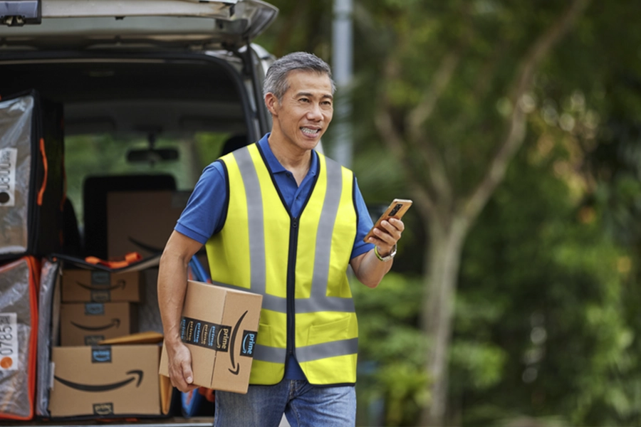 Man in a yellow vest holding a phone and an Amazon package walking away from his vehicle on a tree-lined street